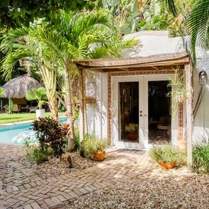 A small white building surrounded by lush greenery and a pool. Tropical plants are in pots, creating a serene, inviting atmosphere.