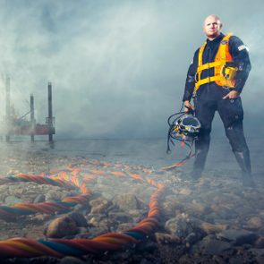 Diver stands on rocky shoreline, holding a helmet. Thick cables lie on the ground, while a platform is visible in the misty background.