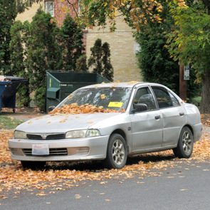 Car covered in autumn leaves, parked on street with recycling bins and trees in residential area; yellow paper visible on windshield.