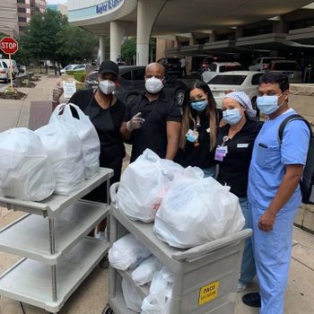 People in masks stand by carts filled with bagged items outside a hospital entrance, showing support. Signs and trees are visible in the background.