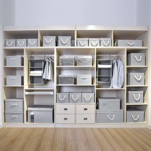 Shelves hold gray storage boxes and some hanging clothes, organized neatly, in a wooden closet against a white wall and a wooden floor.