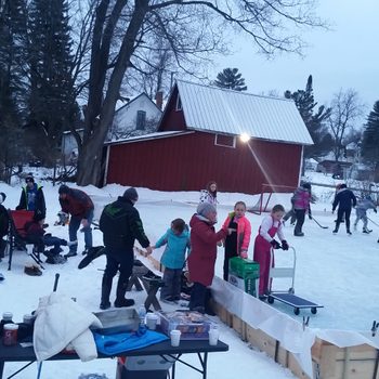 People gather on a snowy ice rink; children play hockey while adults converse by a table with hot drinks. A red barn and trees surround.