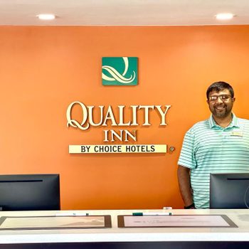 A man stands smiling behind a hotel reception desk with "Quality Inn by Choice Hotels" signage on an orange wall.