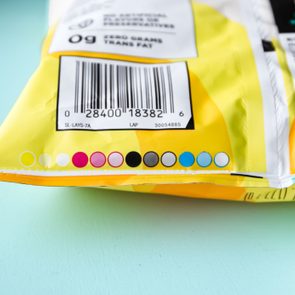Yellow snack bag displays a barcode with colorful circles underneath, resting on a light blue surface. Text includes: 