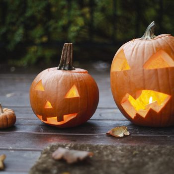 Pumpkins glow with carved faces on a wooden porch, surrounded by small pumpkins and dry leaves, near a brick wall.