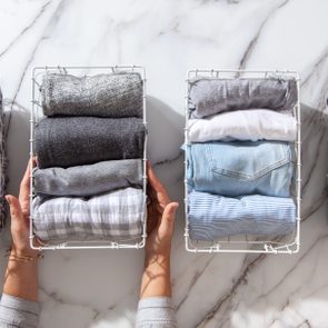 Hands hold wire baskets with neatly folded clothes, set on a white marble surface.