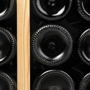 Bottle bottoms are stacked horizontally, their glossy surfaces reflecting light, separated by a wooden divider in a dimly lit storage setting.
