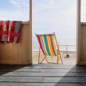 A striped deck chair faces the sea, framed by an open doorway on a wooden deck, with colorful towels hanging nearby.