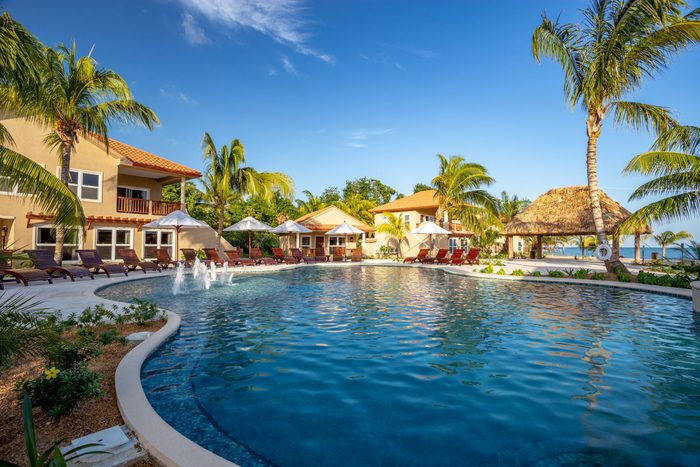 A pool reflects sunlight, surrounded by lounge chairs and umbrellas, with palm trees and a thatched-roof gazebo near tan buildings under a clear sky.
