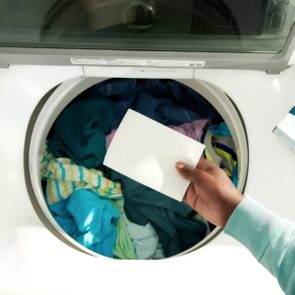 A hand inserts a white sheet into a top-loading washing machine filled with colorful clothes.