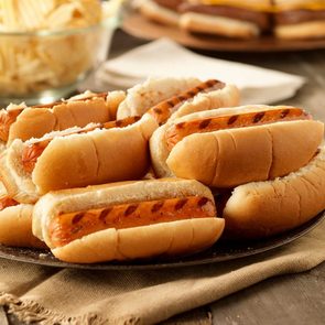 Grilled hot dogs rest on a plate atop a wooden table, accompanied by a bowl of potato chips in the background.