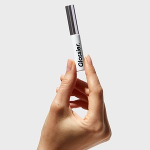 Hand holds a white Glossier tube upright against a plain gray background.