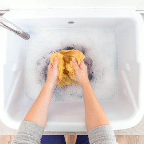 Hands wash yellow fabric in soapy water within a white basin on a light countertop.