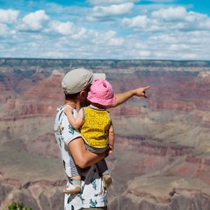 An adult holds a child, both facing and pointing toward a vast canyon landscape under a partly cloudy sky.