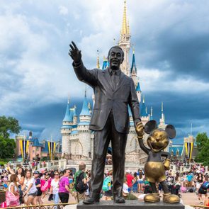 Bronze statue depicts a man holding hands with a mouse character, set against a castle backdrop and surrounded by crowds under a cloudy sky.