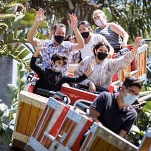 People ride a roller coaster, excitedly raising their arms, surrounded by lush palm trees and vibrant greenery beneath a clear blue sky.