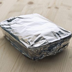 Rectangular aluminum foil-wrapped package sits on a light wooden table, reflecting light softly in a calm environment.