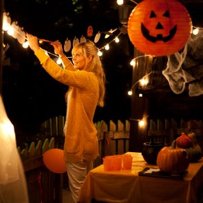 Woman decorates porch with Halloween lights; orange pumpkin lanterns and table with festive items create a spooky, illuminated atmosphere.