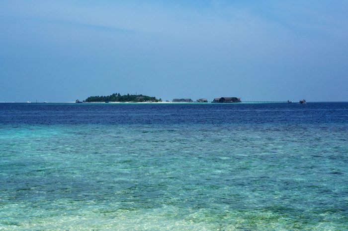 Island sits with small structures on stilts; calm ocean waters surround under a clear blue sky.