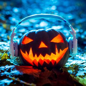 A carved pumpkin wears headphones, glowing brightly amidst fallen leaves, set against a blurred blue-toned background.