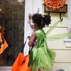 Child in fairy costume, knocking on door, holding a Halloween bag. Another child dressed as a lion, both on a decorated porch.