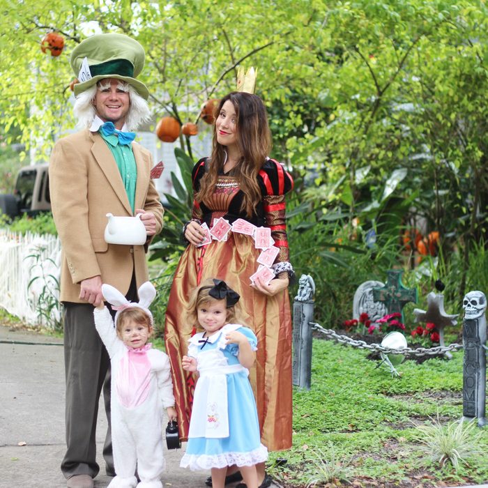 Four people wear elaborate, themed costumes resembling storybook characters, standing on a walkway with green foliage and Halloween decorations around them.
