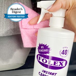 Hand holds Folex carpet cleaner spray bottle and pink cloth, positioned near gray fabric surface. Reader's Digest 