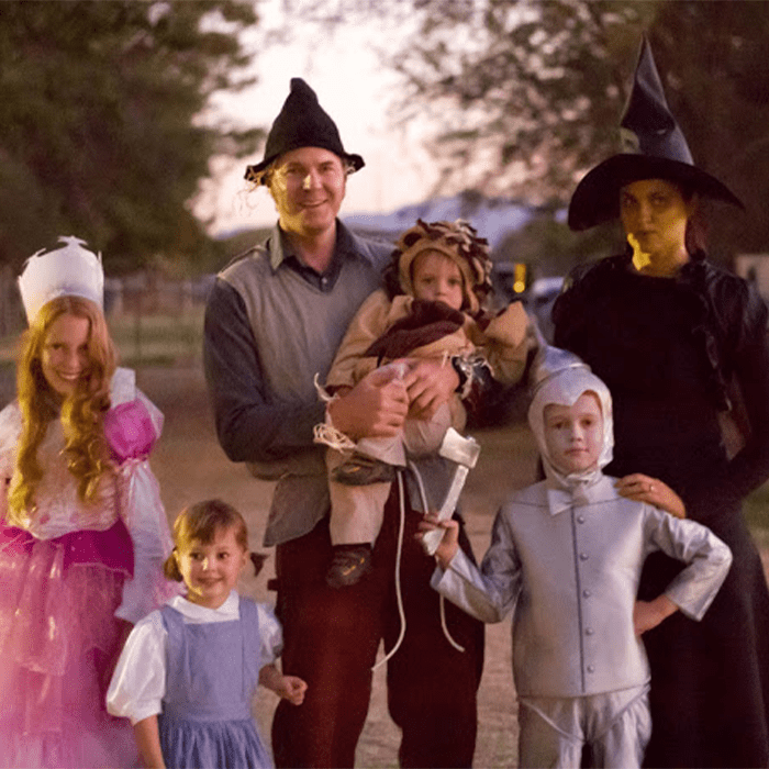 Family wearing character costumes stands outdoors at dusk, gathered together, surrounded by trees and an open field.