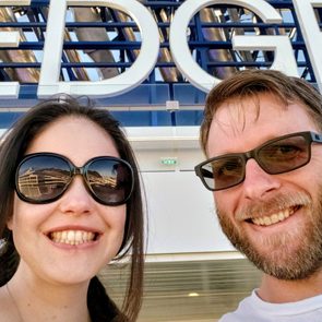 Two smiling people wearing sunglasses pose outdoors against a large 