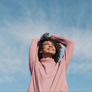 Person smiles, raising arms overhead, wearing a pink sweater against a blue sky backdrop.