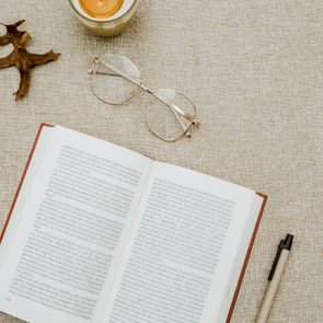 An open book rests on a cloth surface, surrounded by a pen, glasses, small pumpkins, leaves, and a lit candle.