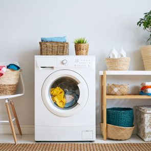 Washing machine operating, surrounded by baskets with laundry, plants, and a shelf with folded clothes in a tidy laundry room.