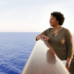 A person leans on a railing, gazing at the ocean, with wicker chairs and cushions nearby, under a clear sky.