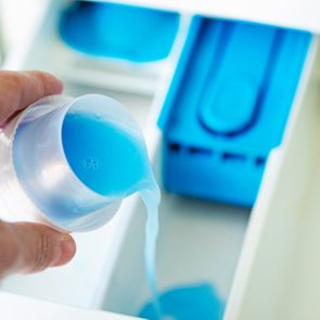 Hand pouring blue liquid detergent into a washing machine drawer, with compartments visible in a bright setting.