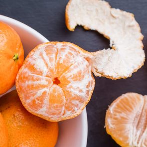 Peeled mandarin orange sits in a bowl with unpeeled ones; segments and peel rest on a dark surface nearby.