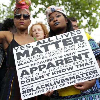 People hold protest signs emphasizing focus on Black lives amid others; outdoors amid a crowd under trees.
