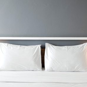 A neatly made bed features two white pillows, a wooden headboard, and stands between two lit bedside lamps against a gray wall.