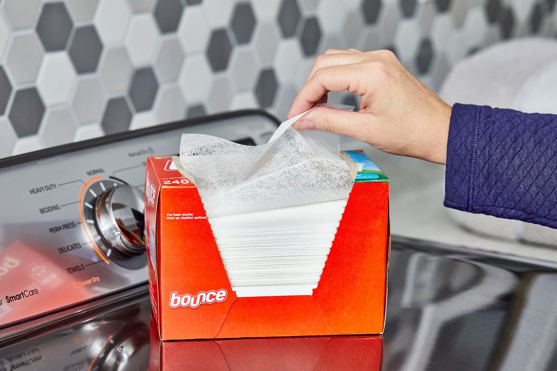 Hand pulls dryer sheet from orange box labeled "Bounce" on laundry machine near hexagonal-tiled wall.