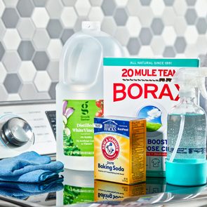 Cleaning supplies arranged on a washing machine, including vinegar, borax, baking soda, and a spray bottle; a hexagonal tiled wall in the background.