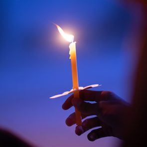 A hand holds a lit candle with a paper drip protector, against a soft blue background.
