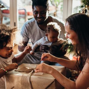 Child excitedly opens a gift with family gathered around, near a decorated Christmas tree in a cozy living room.