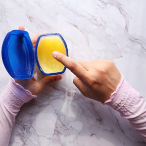 A person's hand dips a finger into a blue container of yellow petroleum jelly on a marble surface.