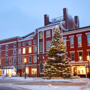 Christmas tree stands decorated with lights and ornaments, surrounded by snow-covered ground and buildings in a town square during evening.