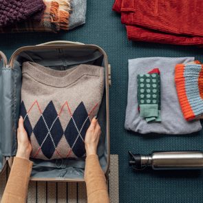 Hands pack a patterned sweater into a suitcase, surrounded by folded clothes, hat, scarf, and a water bottle on a textured surface.