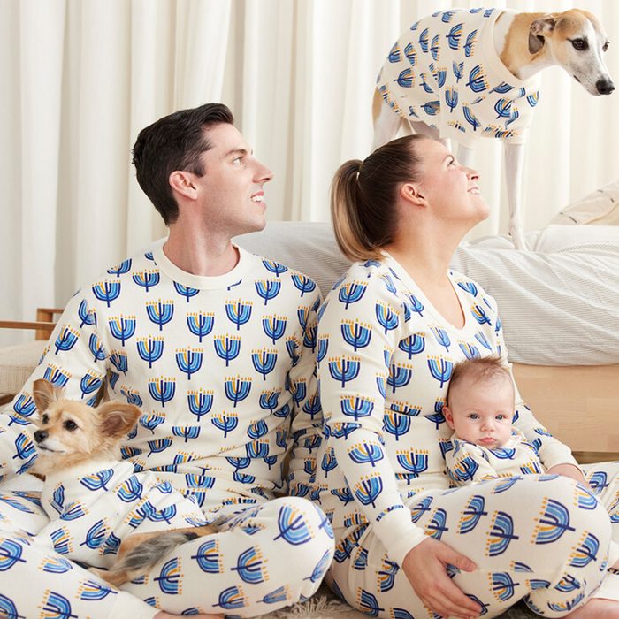 Family and dogs wear matching pajamas with menorah print, sitting together in a cozy living room setting with white curtains.