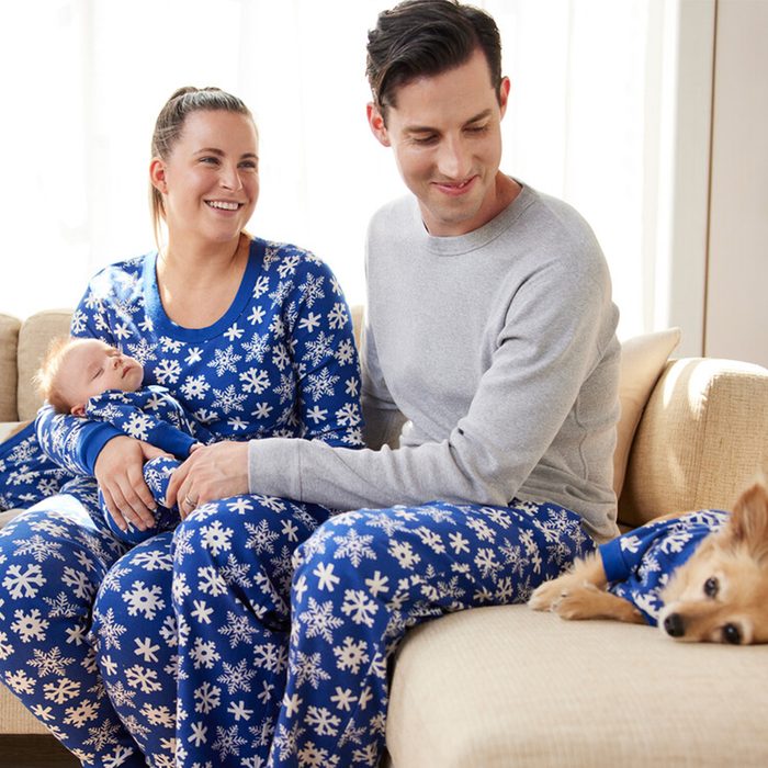 Family wearing matching snowflake pajamas sitting on a couch; baby sleeps in mother's arms, small dog rests beside them in cozy living room.