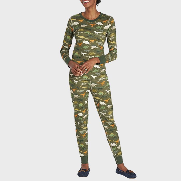 Woman wearing dinosaur-patterned pajamas stands smiling against a plain background, wearing dark slippers.