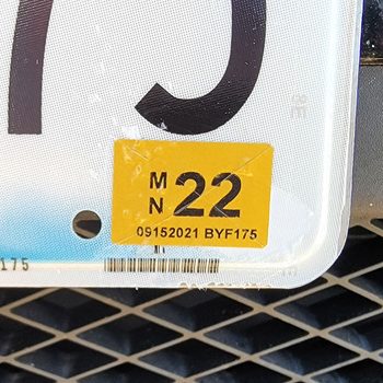 License plate displays a yellow sticker with "MN 22" and "09152021 BYF175," mounted on a vehicle with a grid-patterned grille background.