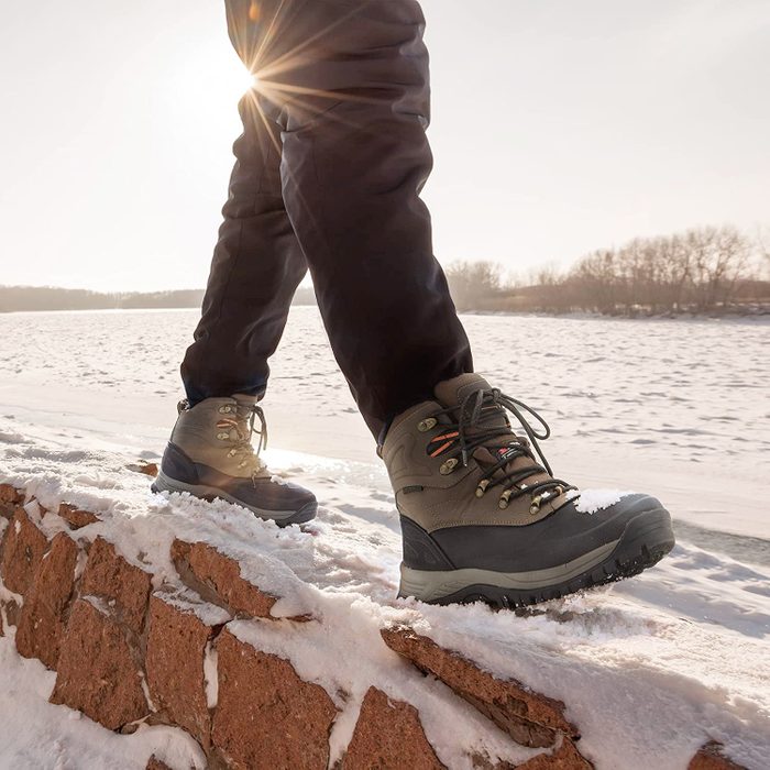Boots walk on snow-covered stone wall by a frozen river, with sunlight shining and trees in the distance.