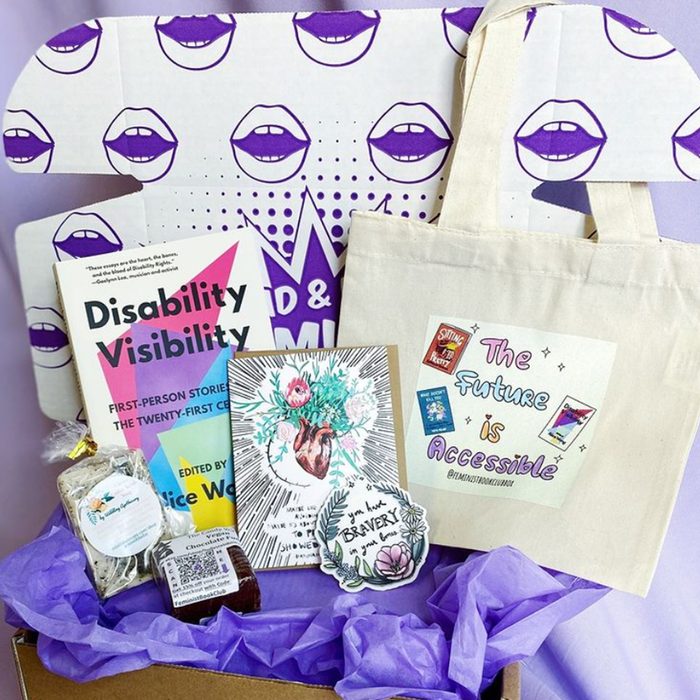 Tote bag holds book, art print, and snacks, surrounded by purple tissue paper, within a box decorated with purple lips on a dotted background.